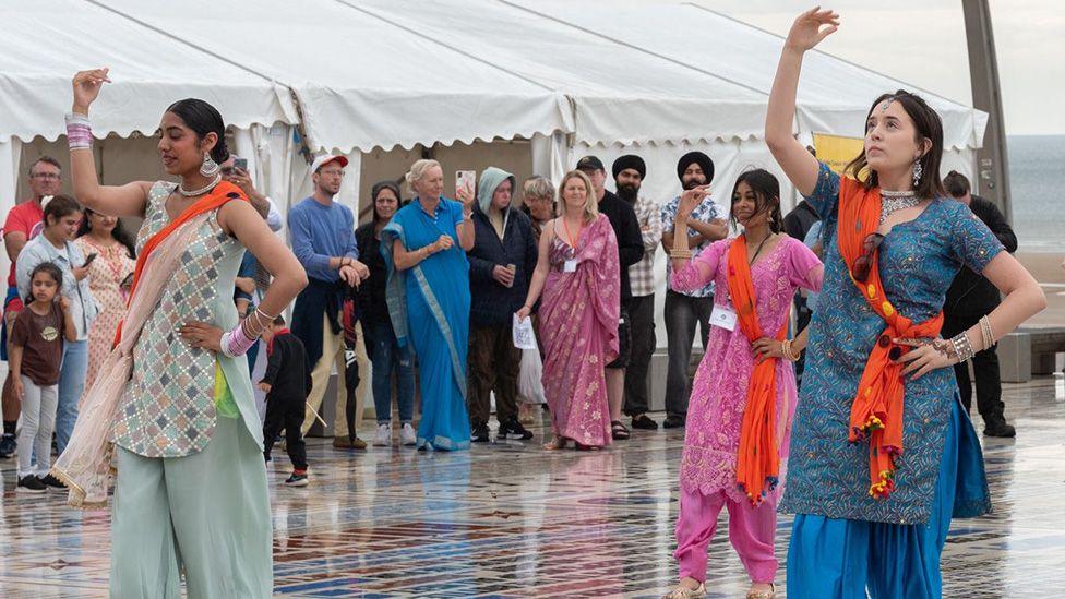 Three young female Indian dancers dancing on the comedy carpet in Blackpool, with their right arms raised and people watching in the background in front of three white marquees.