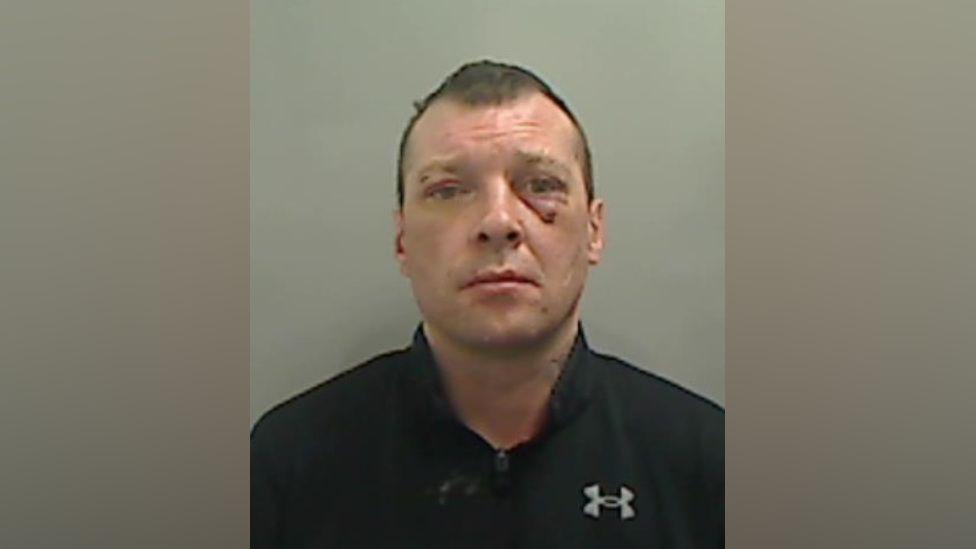 Mugshot of Carl Wild. The image is pixilated but he has several cuts to his face and a black eye. He has short brown hair and is wearing a black t-shirt with the Under Armour logo .