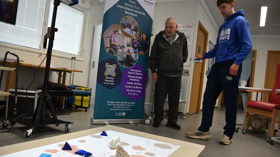 An older and a younger man are standing in front of a sheet spread out on the floor with different items on it. They are in a treatment room.