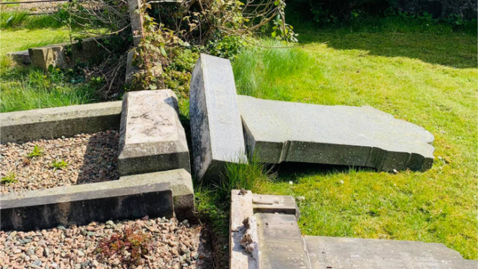 One of the damaged gravestones shown from the side, knocked off its base and lying on the ground