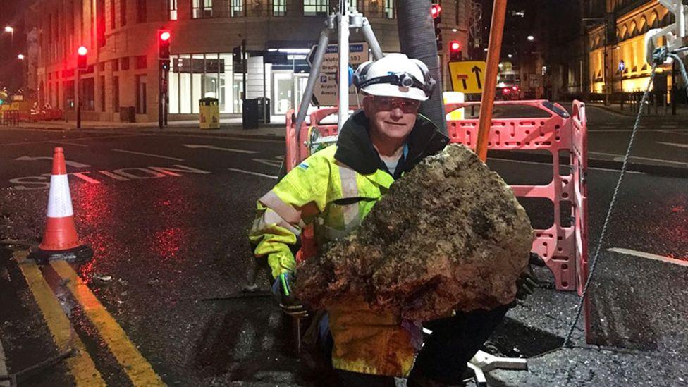 A Yorkshire Water worker in florescent jacket and hard hat emerges from a sewer holding part of a fatberg - it's a large chunk of solidified fat.