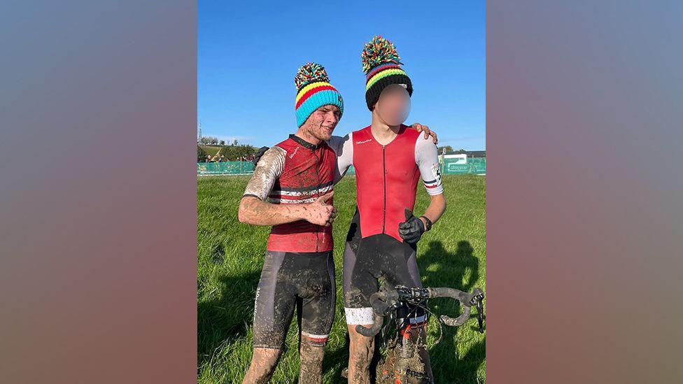 Aidan (l) stands with his arm around another cyclist: two young men, wearing colourful bobble hats, red cycling jerseys and black shorts, stand on the grass and pose for a photograph. They are covered in mud, and the man on the right is straddling a bicylcle.