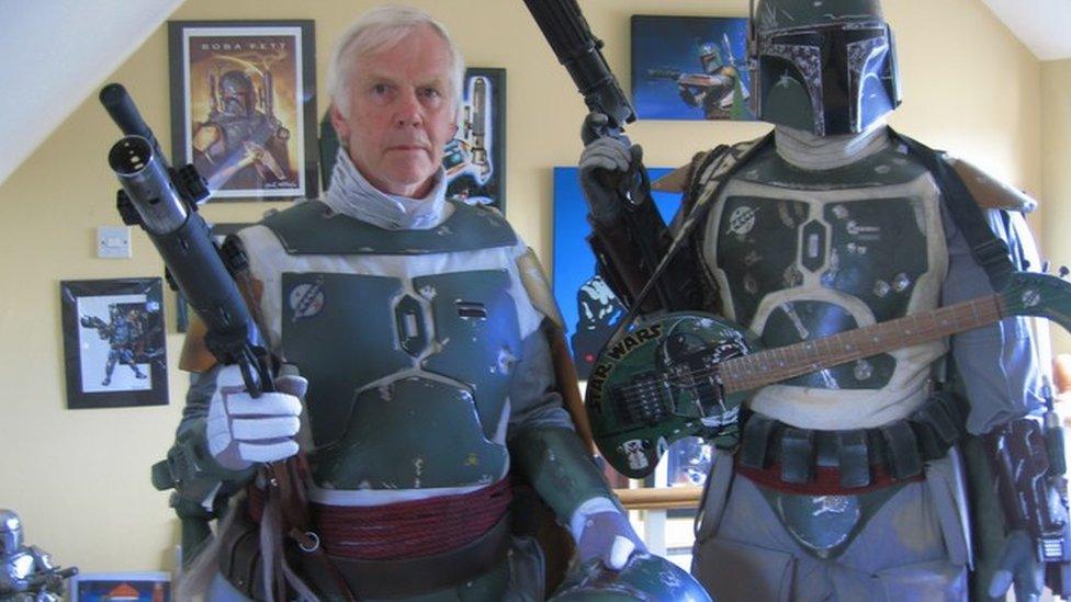 Jeremy Bulloch in Boba Fett costume standing alongside a Boba Fett figure, with Star Wars images on a wall in the background