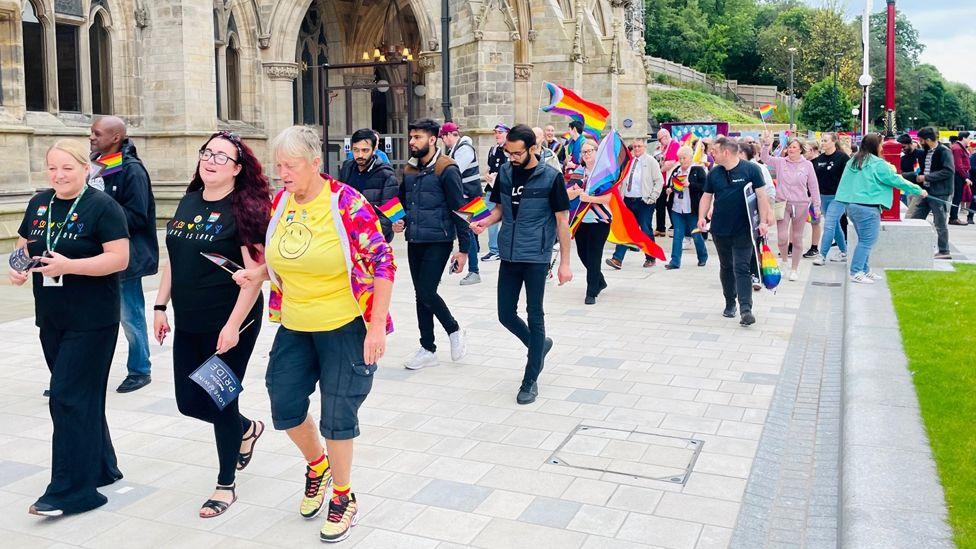 A line of people dressed in colourful clothing and carrying rainbow flags walk in a parade outside Rochdale's stone-built gothic town hall