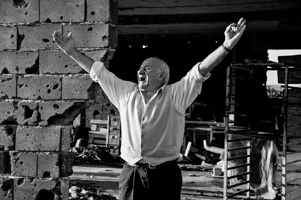 An elderly man holds his hands aloft as he stands next to a war torn building