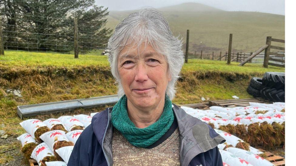 Jenny Wong yn yr awyr agored mewn ardal wledig - mae pecynnau mwsogl ar y llawr y tu ôl iddi