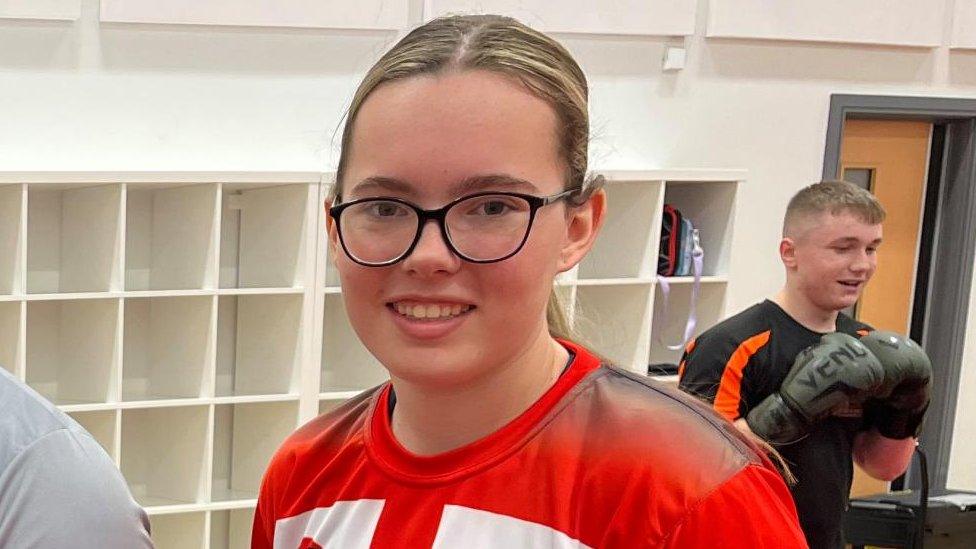 Girl with long hair and glasses wearing a red t-shirt. A man with large boxing gloves is visible in the background.