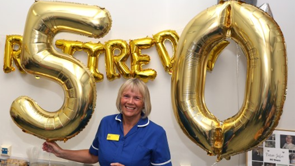 Julie is wearing her uniform and is stood in front of balloon letters spelling out that she has retired as she holds the numbers 5 and 0  in front of cakes and various treats.