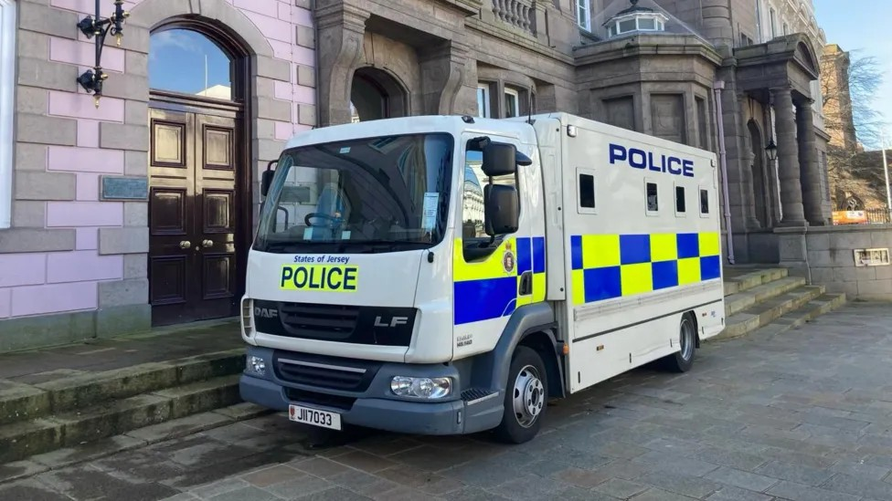 A large police truck parked outside a stone building - it has yellow and blue checked squares and blue lettering