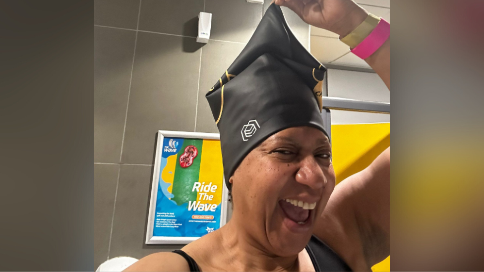 Trish Adudu is taking a selfie while in her swimming costume and wearing a black soul cap on her head. She is looking joyfully at the camera with a open mouth smile. She has one had pulling at the top of the hat to show how much room there is in it for her hair. 