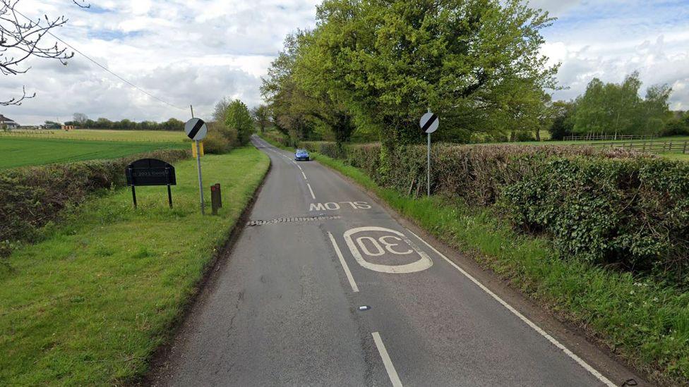 Sarratt Road as it exits Sarratt village. The back of the village sign, which is next to a speed limit sign of a white circle with black diagonal stripe. An identical sign is on the opposite side. There are grass verges and hedges on both sides. Painted in white on the road - upside down from this perspective - is the word SLOW and the number 30 inside a white circle. One blue car drives towards us.