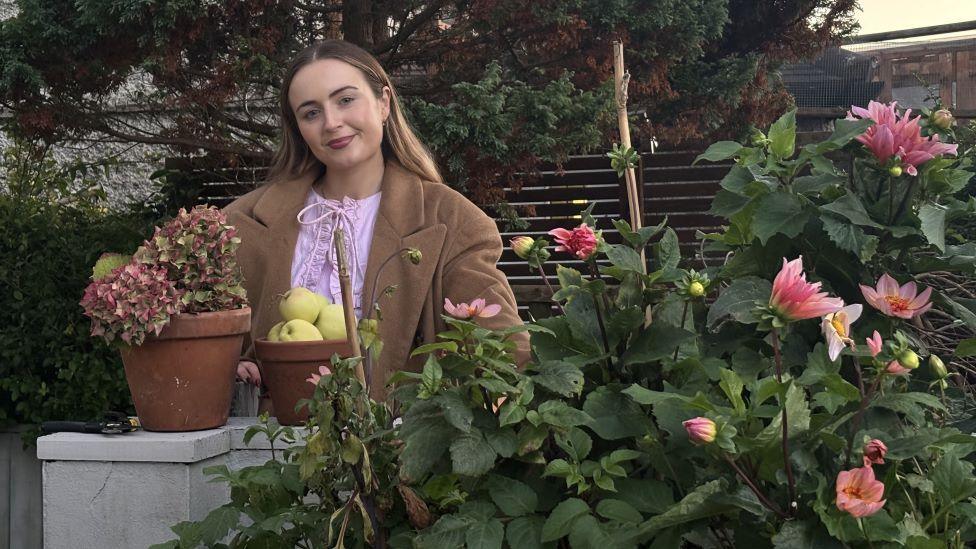 A woman with long brown hair in a brown coat and pink blouse is stood outside behind a plant with pink flowers and in front of a brown fence. She is smiling at a camera and is stood by a plant pot of apples