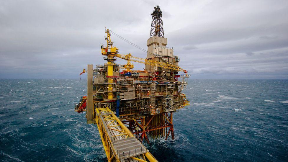 An oil rig in choppy blue waters in the North Sea. The structure is made up of yellow, red and blue pipes and beams, with a tower on the right hand side extending into a grey sky