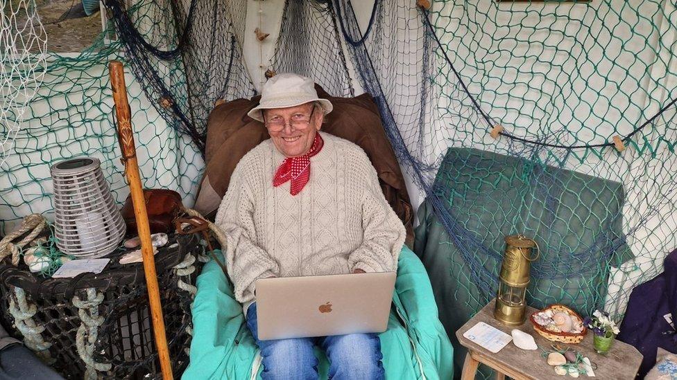 Peter Butler smiling at the camera while sitting in an armchair in a converted shed at the bottom of his garden. He is wearing a white bucket hat, glasses resting on the tip of his nose, a cream knitted jumper, jeans and a red handkerchief around his neck. There is a laptop resting on his knees. He is surrounded by aquatic-themed decorations, such as lobster pots, ropes, nets, shells and a gold ship lantern.