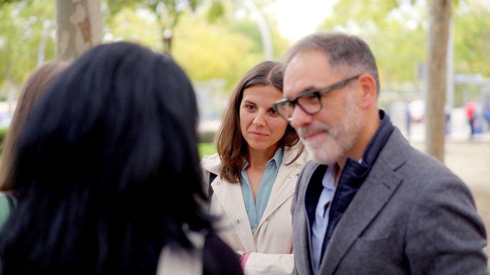 Victoria (seen only from the rear) with thick dark hair, stands in the park talking to Cristina and Felix 