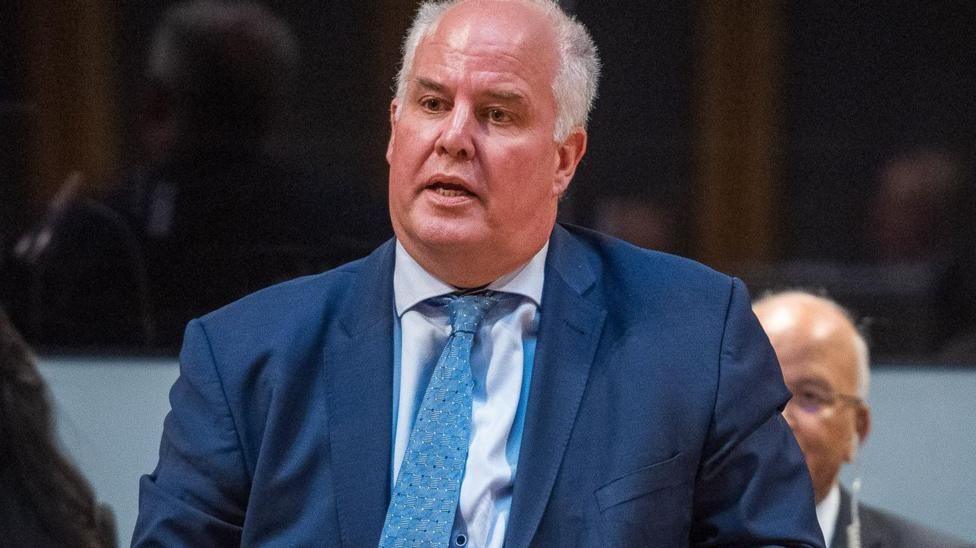 Andrew RT Davies speaking in the Senedd chamber wearing a blue suit jacket, light shirt and a tie. The face of another Conservative politician can be partly seen behind him.