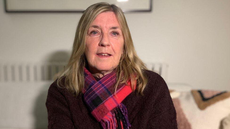 A woman with blonde hair, wearing a maroon jumper and red tartan scarf, looks straight ahead in a close-up shot 