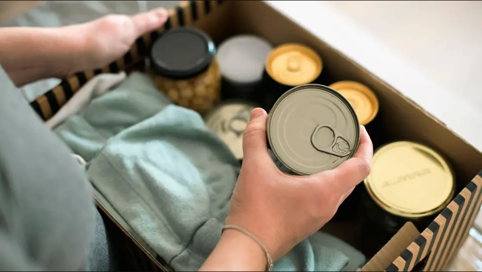 A close-up of a box of tinned food. A person is holding the box. They are clasping one of the tins in their right hand.