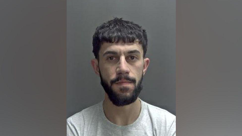 Safa Lawan is looking at the camera in a police custody image. He has dark, short hair and a dark beard and moustache. He is wearing a grey T-shirt