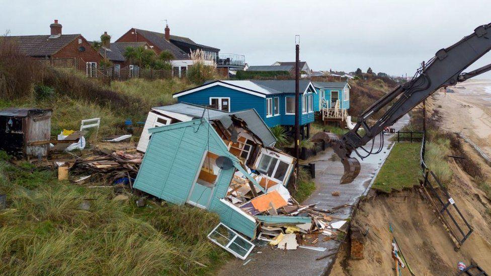 A large piece of a machinery tearing down a light blue wooden chalet, which lies just a few feet from a cliff edge. Other chalets line the cliff edge.