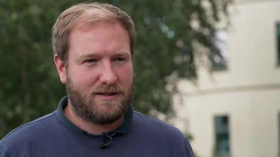 Paul Miller wearing a blue jumper. He has brown hair and a brown beard. The camera is focused on him, but behind, you can see a blurred tree and a white building on the right.