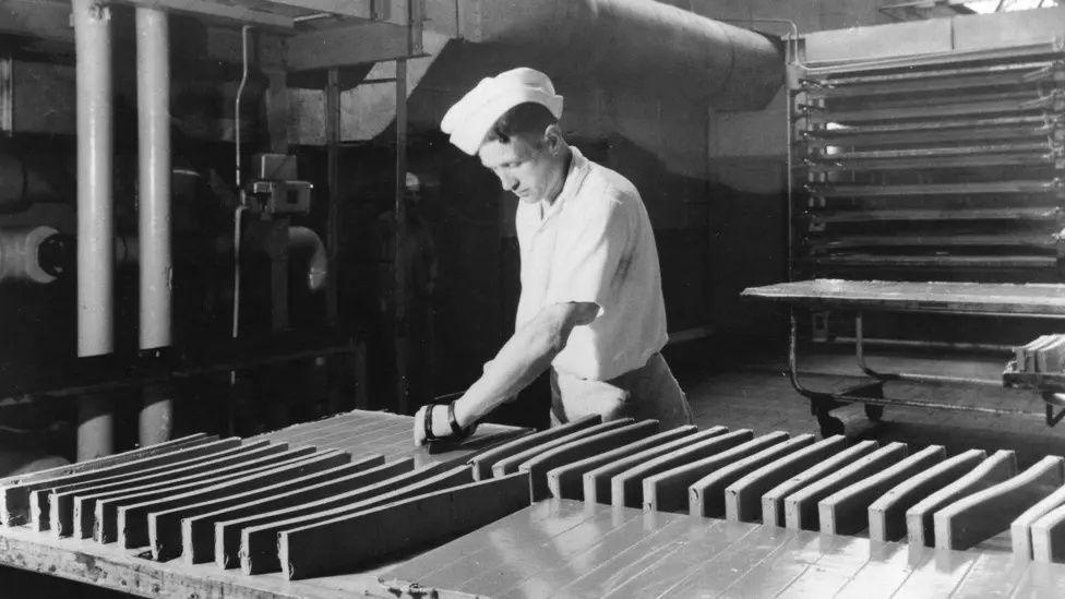 A black and white photo of a man wearing a white shirt and a white hat cutting large slabs of chocolate on a table.