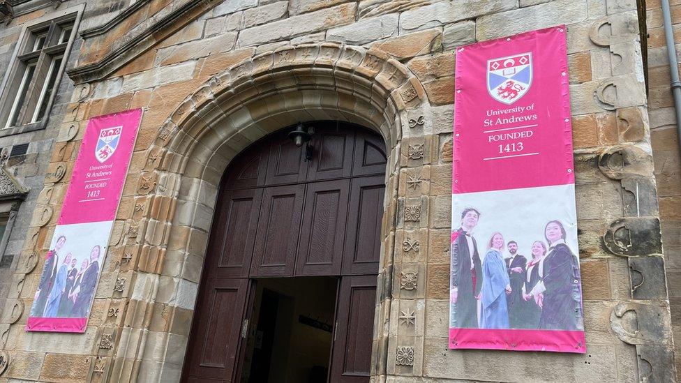 A grand wooden door at the front of an old building at St Andrews is flanked by two flags illustrating the founding of St Andrews in 1413