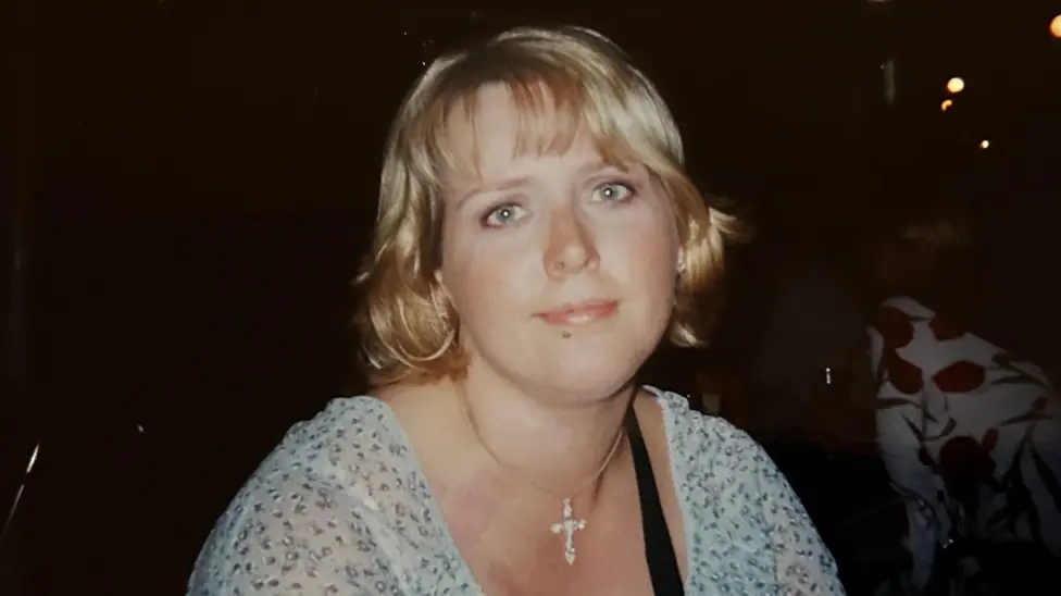 A picture of Clare Wood, who is wearing a light blue top, a crucifix around her neck and has blonde hair. She is looking at the camera.