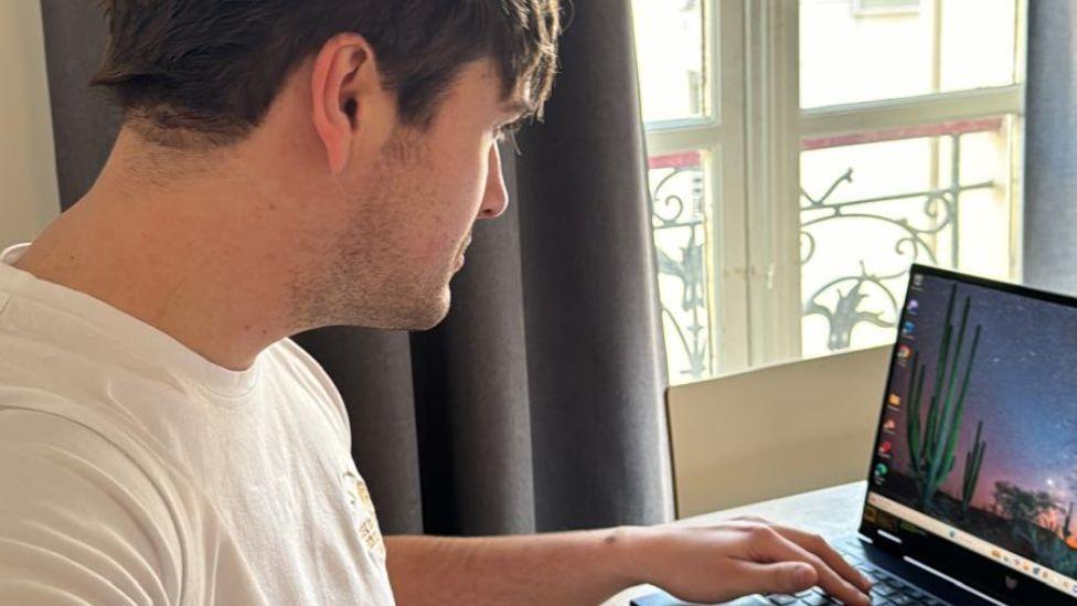 Osian looks at his laptop. He is wearing a plain white t-shirt and he has stubble, with his dark hair cut short. His laptop desktop shows an image of the desert sky at night, with cacti in the foreground. 