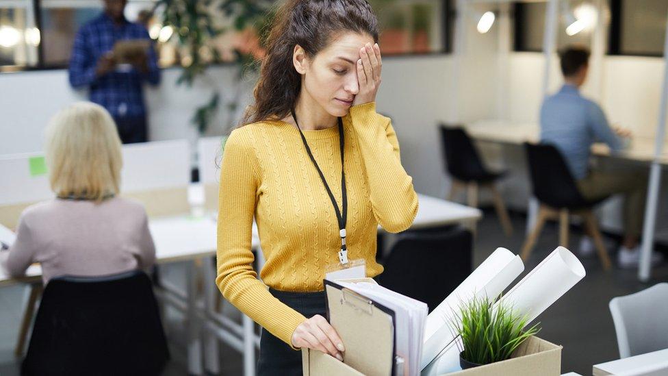 A woman with hand on her face looking worried. She has a cardboard box with a pot plant, books and posters. She is in an office.