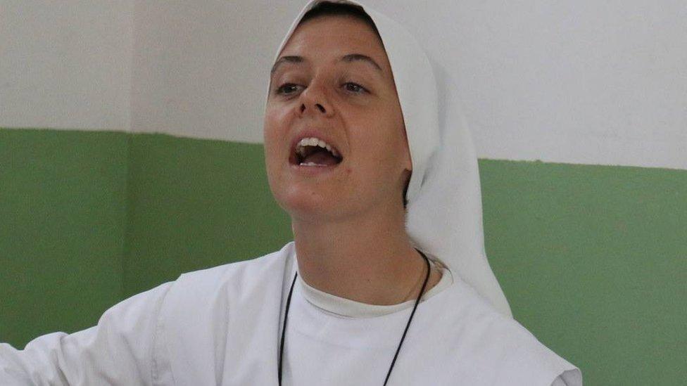 Sr Clare Crockett in white habit, appearing to be singing. she is sat against a green and white wall