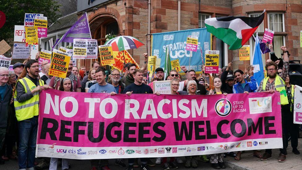 Dozens of protesters line up behind a large pink "refugees welcome" sign. It is held up by the front row, with others holding signs above their heads. They are standing in front of a stone building.