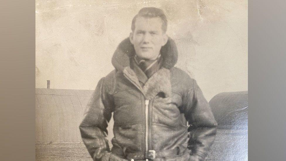 A black and white 1940s or 1950s photo of George Kelly. He is in uniform, wearing a leather flying jacket done up with a zip and with its fur collar raised around his neck. His hands are in his pockets. Behind him can be seen corrugated Nissan huts.