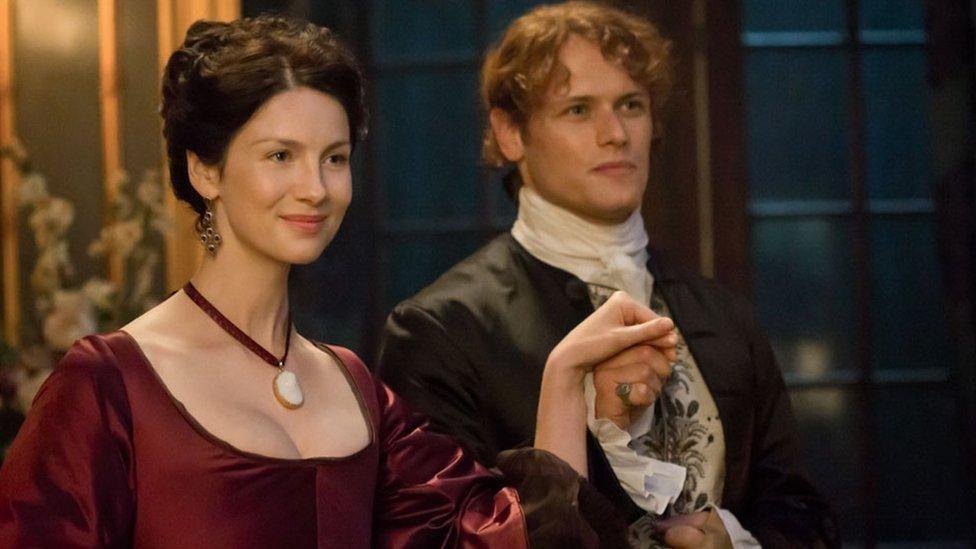 Caitríona Balfe and Sam Heughan in a scene from Outlander - she is dressed in a red dress and has dark hair, while he is dressed formally. They are holding hands and smiling, and appear to be about to take to the dance floor.