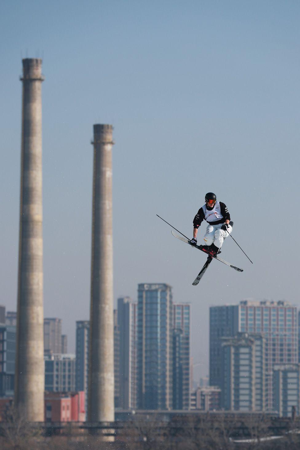 A freestyle skier performing a mid-air trick against a backdrop of a city skyline and clear blue grey sky.