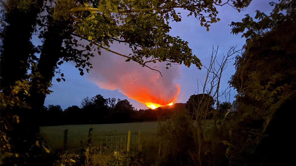 The scene at dusk from a further distance away shows fields and trees in the foreground with an orange glow on a hillside in the distance