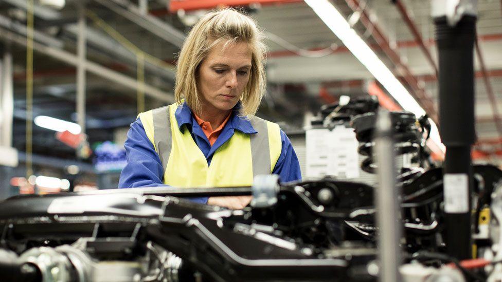 A woman with blonde hair wearing a yellow high-vis jacket and blue shirt works on machinery in a factory