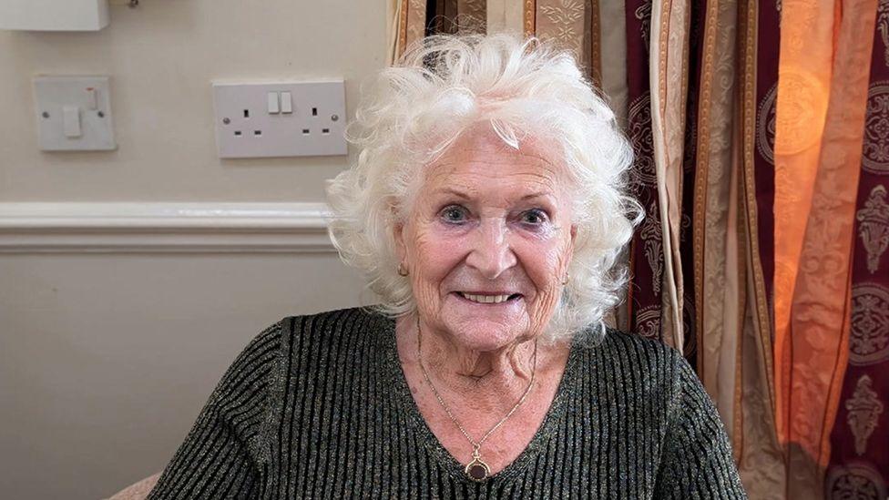 Jackie Holden sat in a chair wearing a grey jumper and a pendant