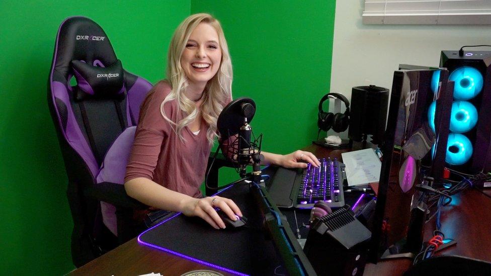 A young blonde woman sits in a black and purple gaming chair in front of a wall covered in green screen material. On the desk in front of her is a professional streaming set-up - a microphone with pop shield, two monitors, a pair of headphones on a stand and a computer terminal with three fans illuminated by light blue LEDs