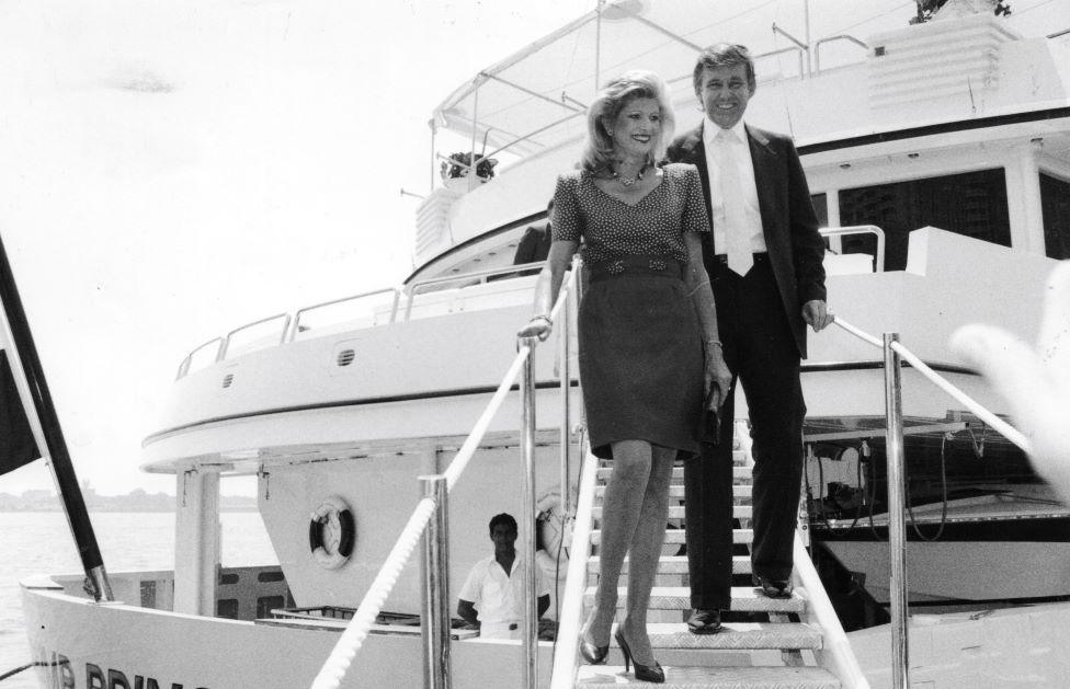 Black and white photo of Donald and Ivana Trump stepping off the Trump Princess yacht. She is wearing a dress and he is wearing a dark suit with a white tie. The yacht is white and the sea is in the background.