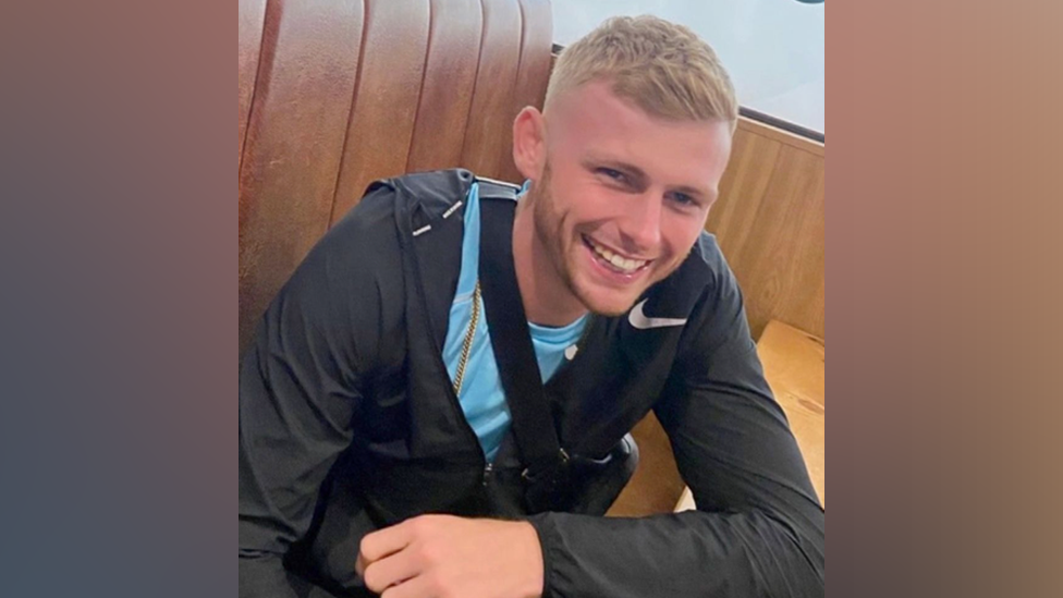 A man with short blonde hair and wearing a black sports jacket and blue shirt, leans on a table with his left arm while smiling.