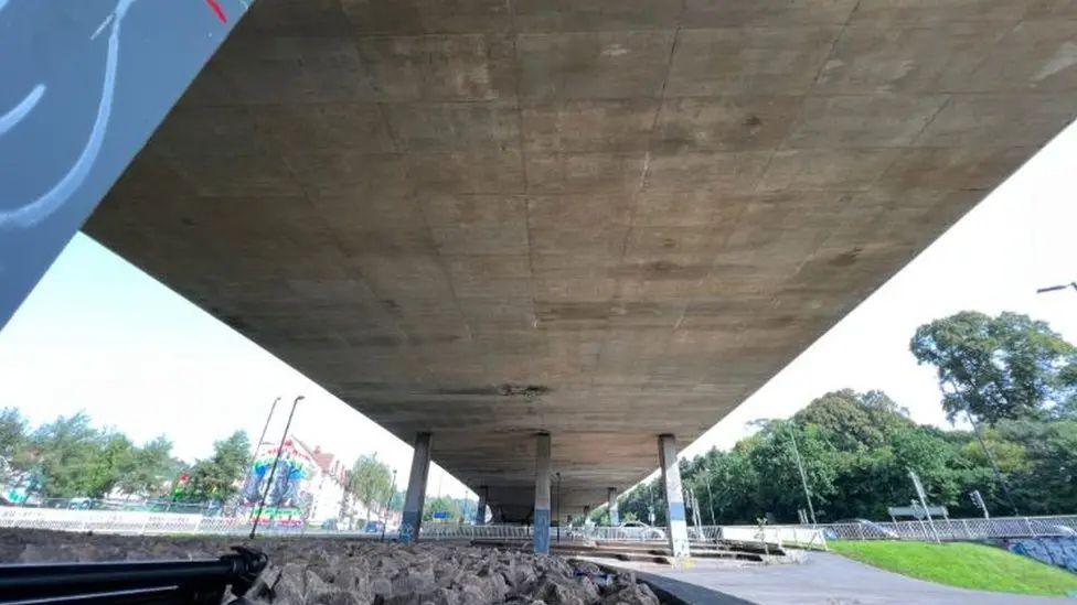 The Eastville Viaduct in Bristol. The image shows the underside of the viaduct. Pillars can be seen in the distance, supporting the structure. Houses and trees can be seen either side of the structure. 