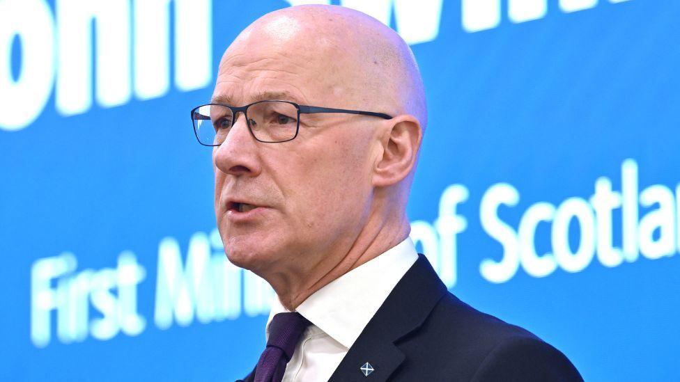 First Minister John Swinney, a bald man with glasses, speaks in front of a blue screen at a podium in a close-up shot 