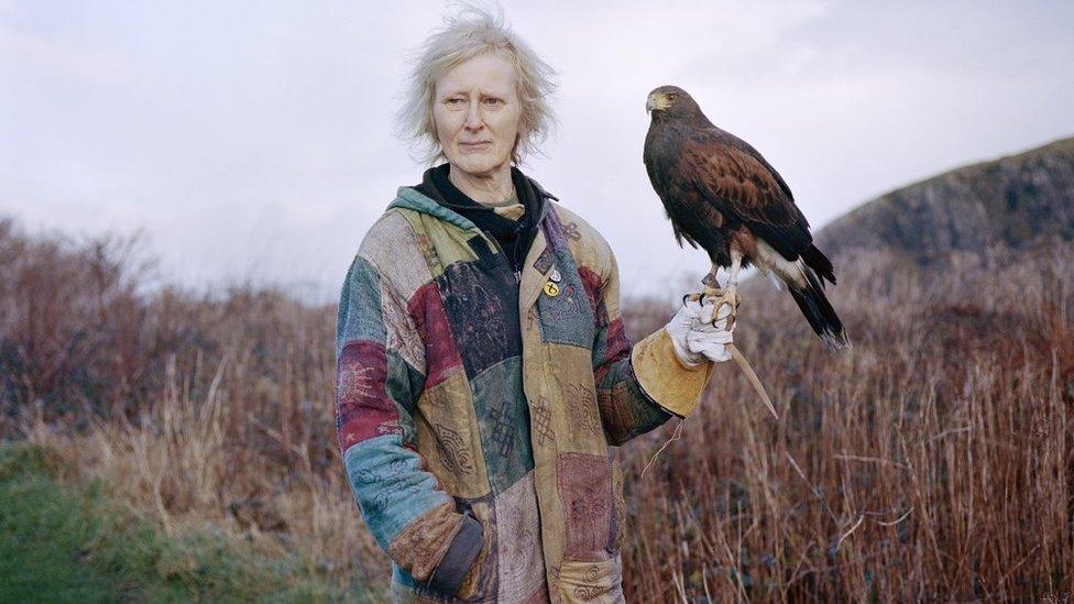 A woman in a colourful jacket stands in the countryside and holds a hawk