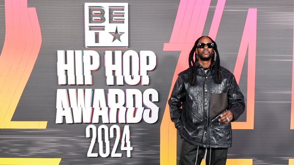 2 Chainz attending the 2024 BET Hip Hop Awards at Drai’s Beach Club – Nightclub in Las Vegas. He is wearing black sunglasses, a shiny black jacket, black trousers and a silver ring on his left hand. He is standing in front of a black and yellow background with the words BET Hip Hop Awards 2024 on it.