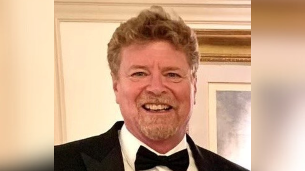 Robert Mabey, who has curly light brown hair and a goatee, smiles at the camera. He is wearing a black suit jacket, white shirt and black bow tie.