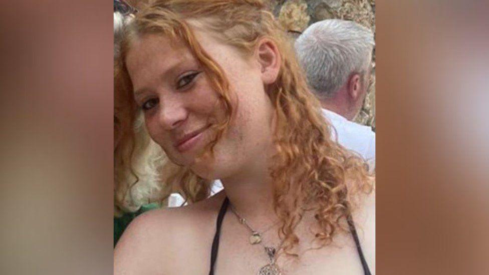 Woman with reddish tight-curled hair she is wearing necklaces and has her head to the left smiling at the camera.