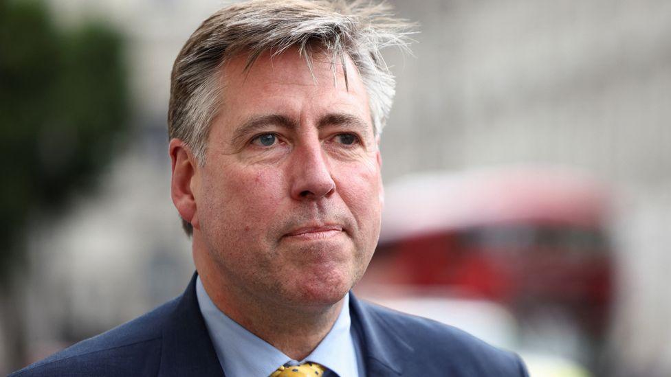 Lord Graham Brady, with brown and grey short side-parted hair and wearing a blue suit, blue shirt and yellow patterned tie, stands in front of a blurred Houses of Parliament and red London bus