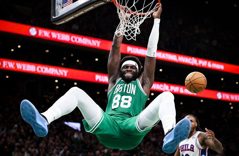 A male basketball player in a green Boston Celtics jersey hangs from the rim after a dunk, legs extended, with another player in a white Philadelphia jersey nearby and a basketball mid-air.