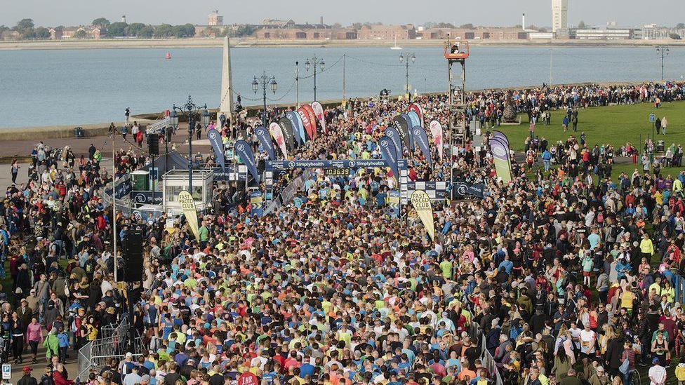 Great South Run setting off from Clarence parade in Portsmouth in 2023. Thousands of runners and spectators can be seen from a bird eye's view. The sea and the city can be seen in the distance. It's a sunny day.